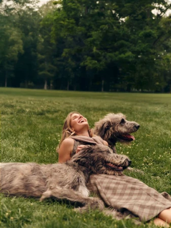 Constance Jablonski laughing while lying on a grassy meadow surrounded by two large Irish wolfhounds.