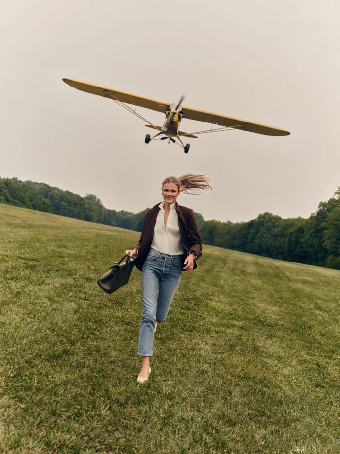 Constance Jablonski running across a grassy field carrying a briefcase as a yellow vintage propeller airplane flies low overhead.