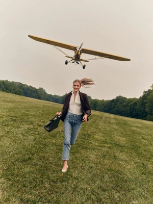 Constance Jablonski running across a grassy field carrying a briefcase as a yellow vintage propeller airplane flies low overhead.
