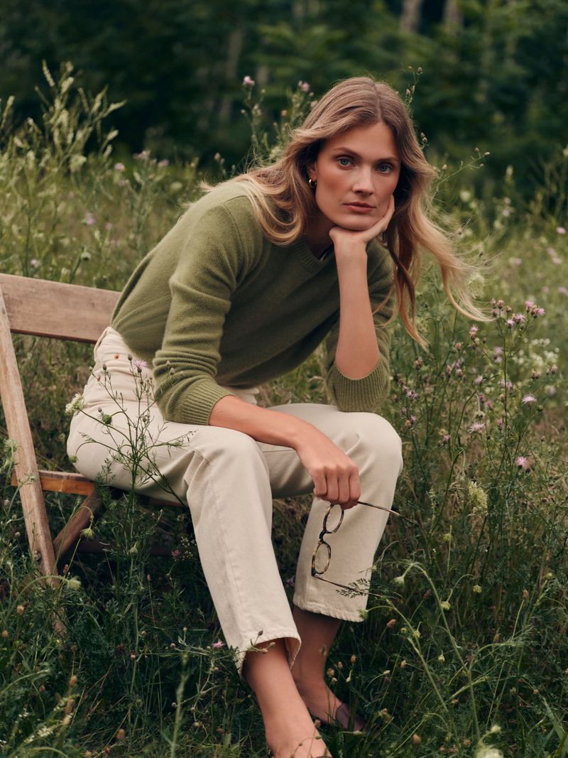 Constance Jablonski seated on a wooden chair in a grassy meadow wearing an olive sweater and cream trousers.