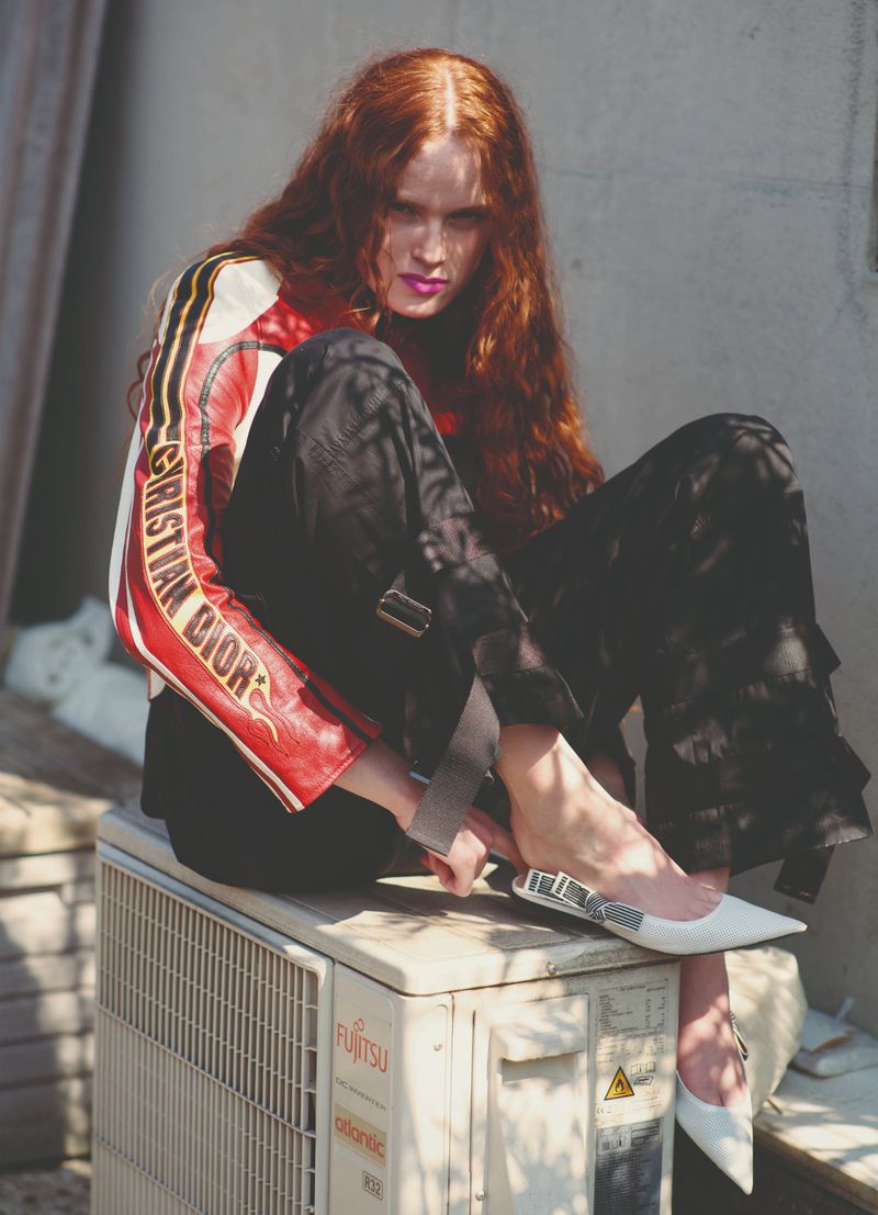 Fashion editorial style outdoor photo of a red-haired woman wearing Dior jacket, black pants and white heels, sitting on air conditioning unit.