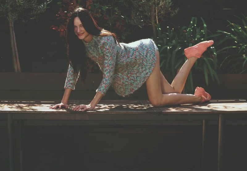 A young woman in a patterned dress and pink shoes poses on a wooden table outdoors, surrounded by green plants in natural lighting.
