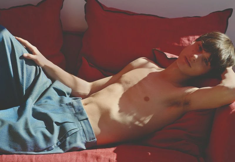 A young adult male with dark hair reclines shirtless on a red couch. He's wearing light blue pants in a brightly lit indoor setting.
