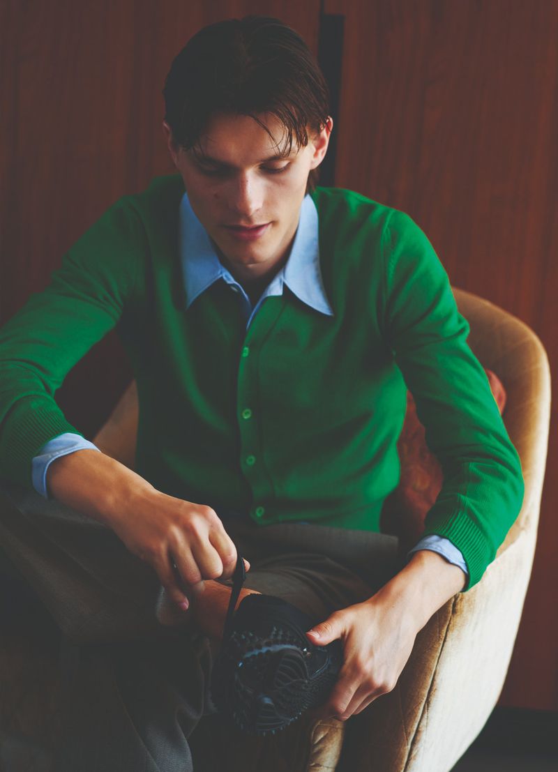 Man wearing a green cardigan and blue collared shirt, seated while polishing a black shoe with a soft cloth indoors, looking down.