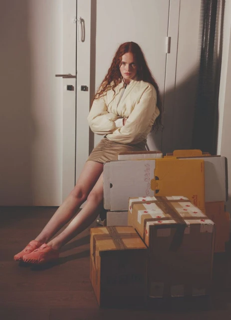 Young woman with red hair sits amongst shipping boxes in front of a white door wearing a light-yellow jacket and a tan miniskirt.