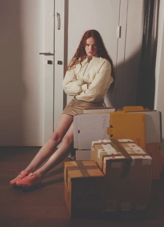 Young woman with red hair sits amongst shipping boxes in front of a white door wearing a light-yellow jacket and a tan miniskirt.