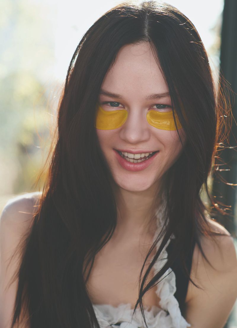Smiling young woman with dark hair wearing gold under-eye patches, indoors, in natural light. Beauty and skincare concept.