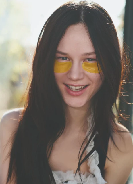 Smiling young woman with dark hair wearing gold under-eye patches, indoors, in natural light. Beauty and skincare concept.
