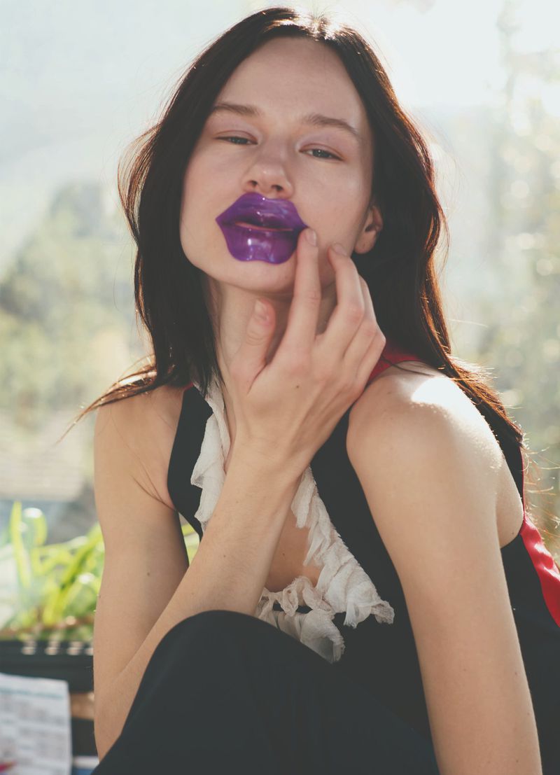 A young adult woman with long dark hair wearing a lip mask and dark clothing poses indoors with a blurred natural background.