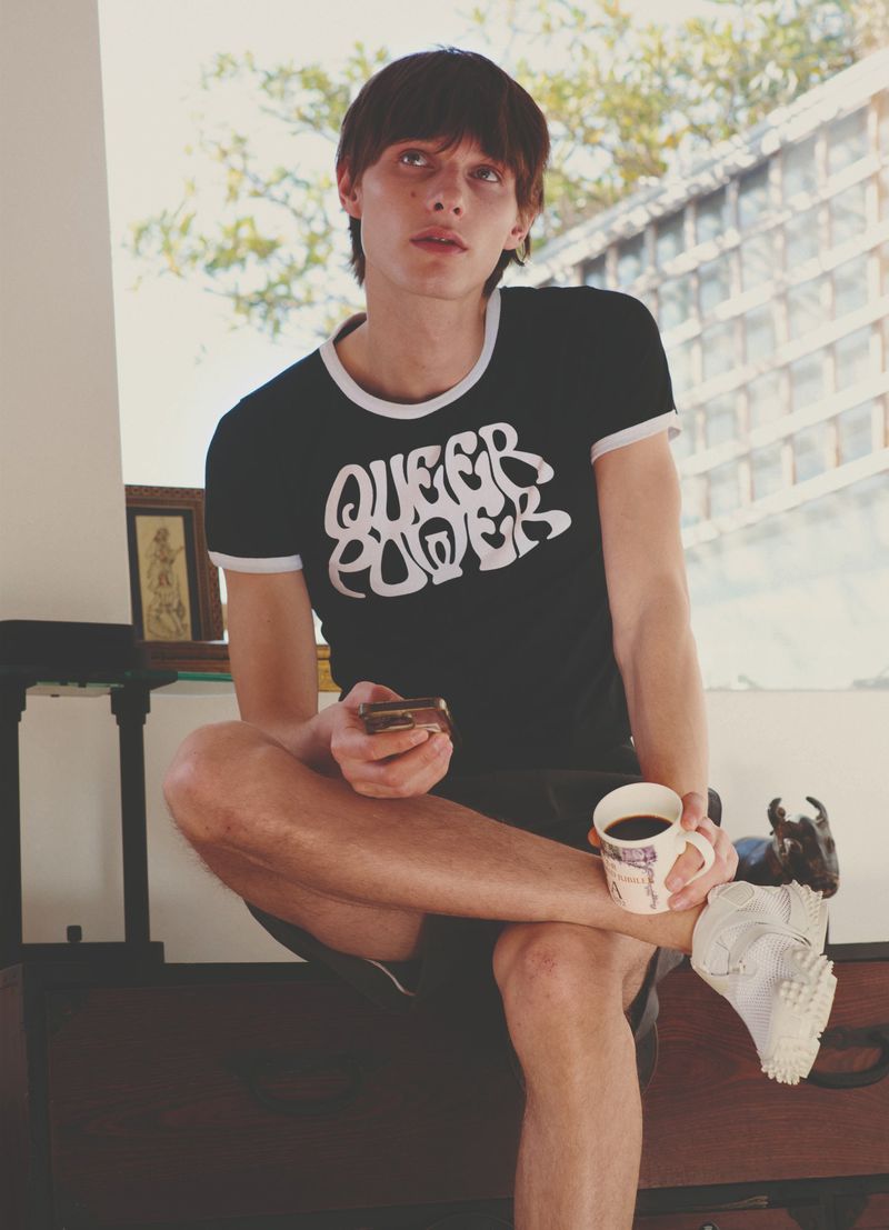A young adult male seated indoors, wearing a black 'Queer Power' t-shirt, holding a coffee cup and a mobile phone, looking off camera.