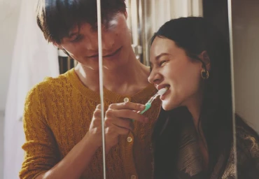 Man in yellow textured cardigan brushing a smiling woman's teeth in a mirror reflection