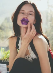 Model with dark hair touching vibrant purple lips wearing a dark sleeveless top against a softly lit natural background.