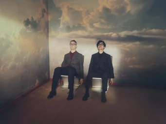 Nicolas Winding Refn and Hideo Kojima seated in tailored suits against a painted cloudy sky backdrop for Prada Mode NYC.