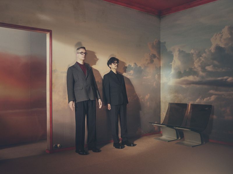 Nicolas Winding Refn and Hideo Kojima standing in dark suits against cloud-painted mural walls in a set-dressed interior for Prada Mode.