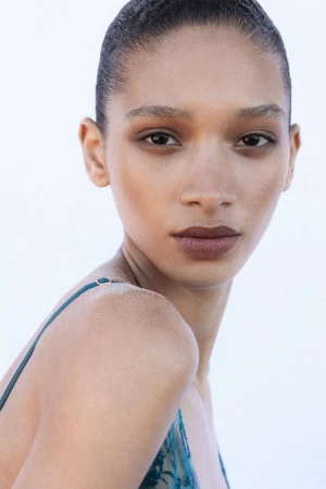 Close-up portrait of model Elodie Guipaud with slicked-back hair and a teal lace strap top against a seamless white background.