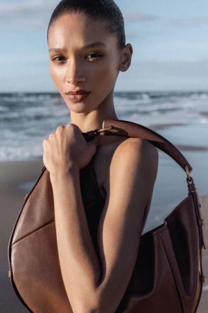 Model Elodie Guipaud holding a curved dark brown leather shoulder bag against an ocean beach background.