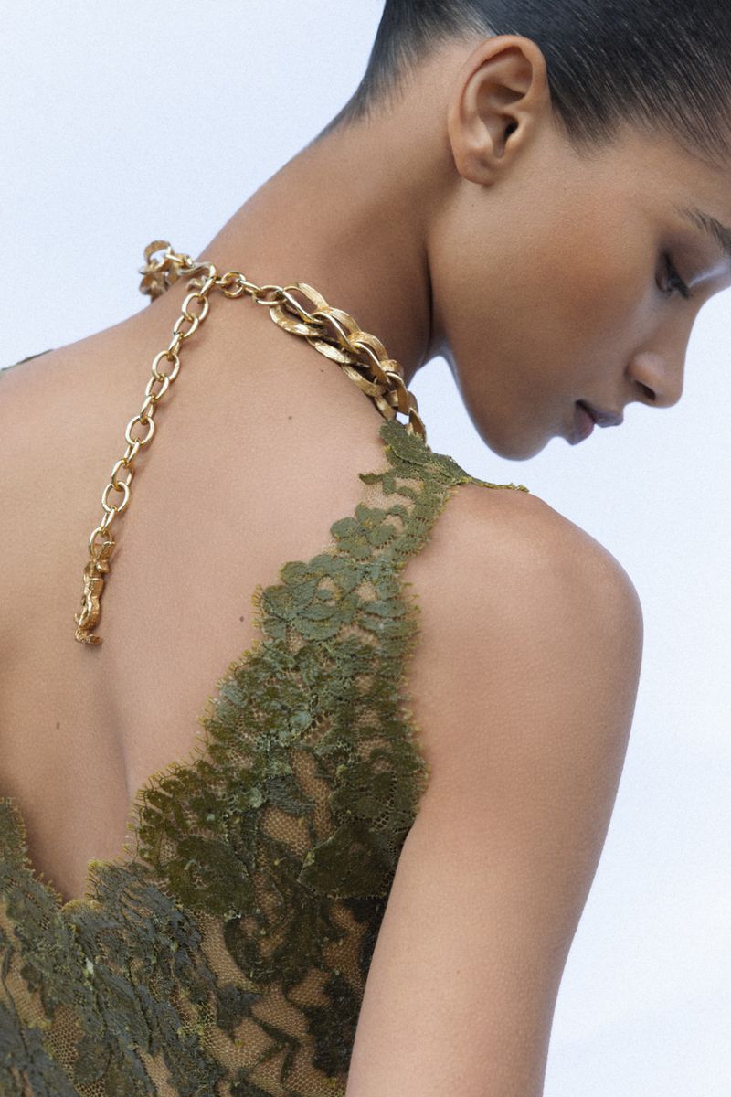 Model Elodie Guipaud seen from behind wearing an olive green lace dress and a gold chain necklace against a white background.