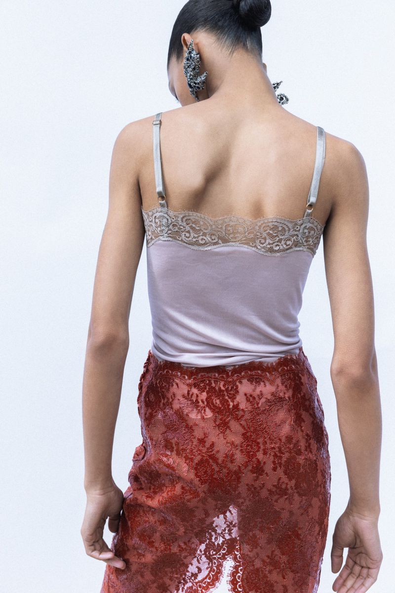 Model seen from the back wearing a pale mauve lace-trimmed camisole top and sheer rust lace skirt against a white backdrop.