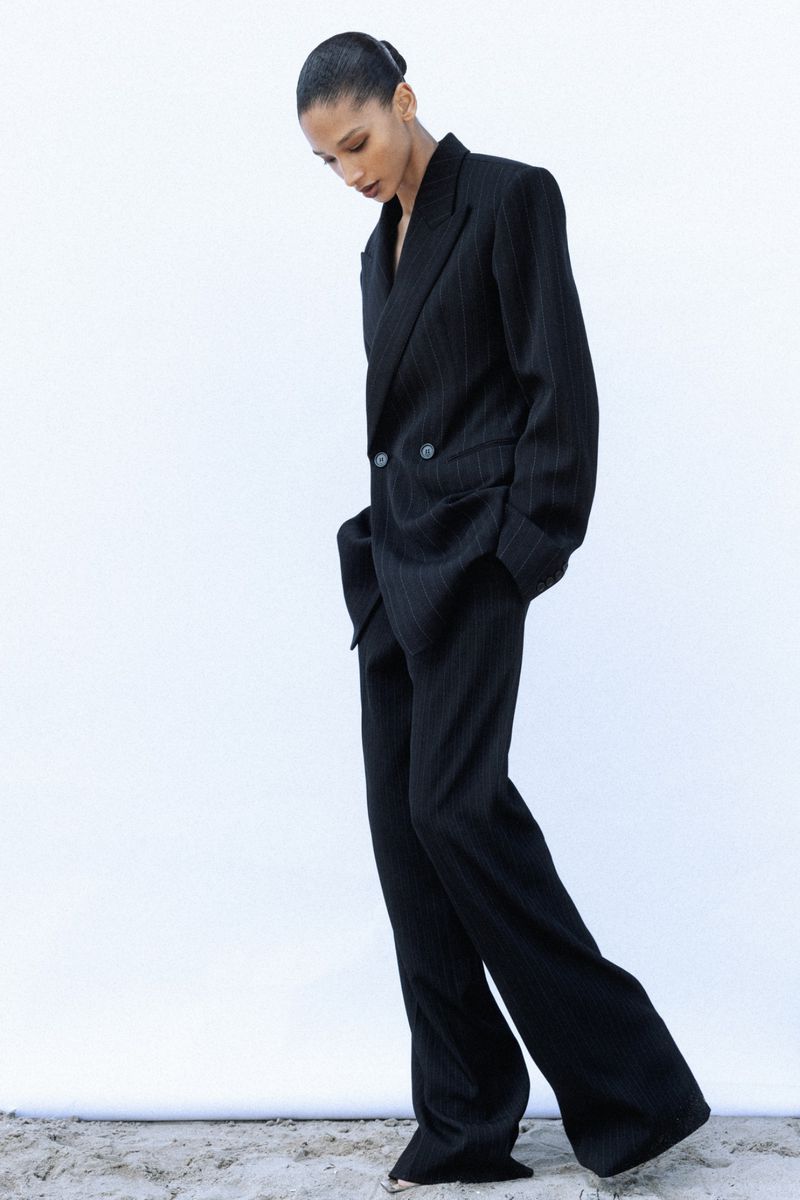 Model wearing an oversized black pinstripe pantsuit standing on sand against a bright white background.
