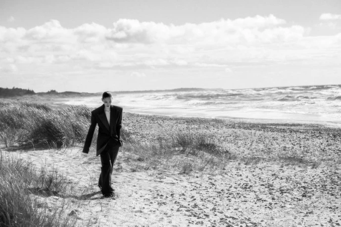Model Elodie Guipaud walks along a grassy sand dune beach wearing an oversized dark tailored suit in black and white.