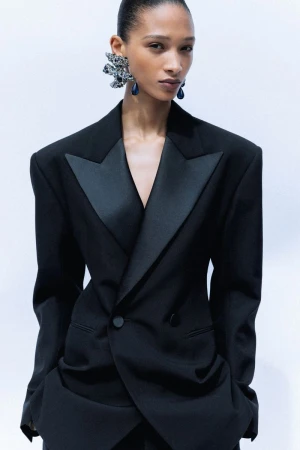 Model wearing an oversized black tuxedo blazer with wide satin lapels and sculptural statement earrings against a plain white backdrop.