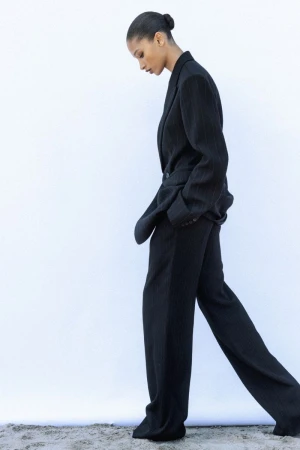 Model Elodie Guipaud in an oversized black tailored suit walking in profile against a bright white backdrop.