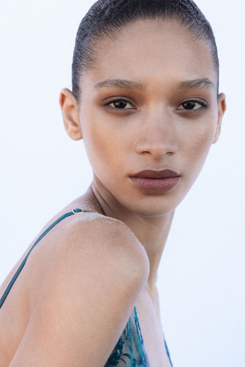 Close-up portrait of model Elodie Guipaud with slicked-back hair and a teal lace strap top against a seamless white background.
