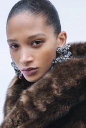 Model Elodie Guipaud wearing a plush brown fur coat and sculptural crystal earrings against a white background.