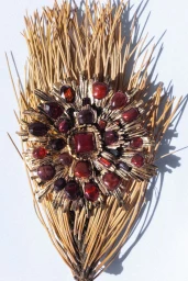 An ornate gold sunburst brooch with red and amber stones resting on dried pine needles over a white surface.
