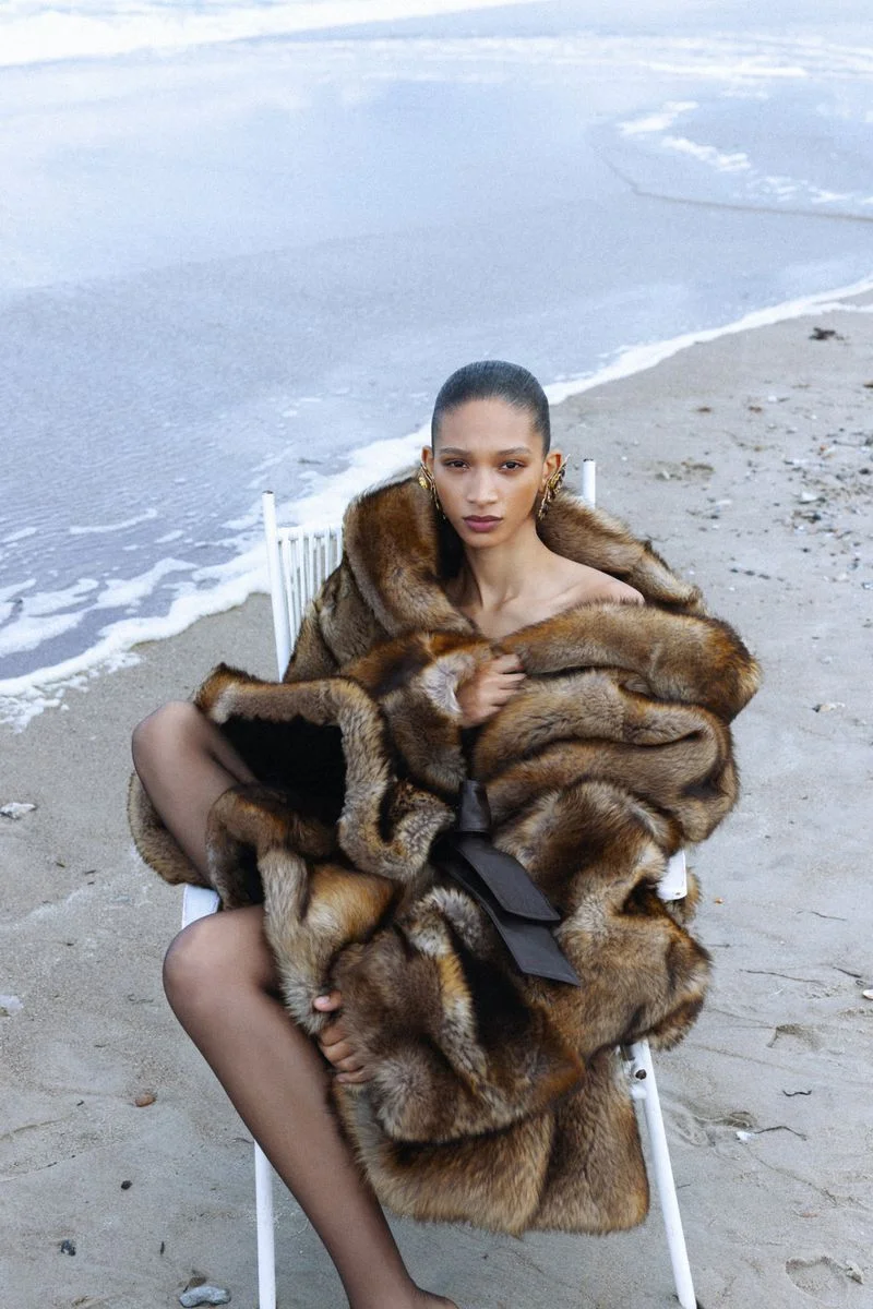 Model Elodie Guipaud seated in a white chair on a sandy beach wearing an oversized brown fur coat with a dark waist tie.