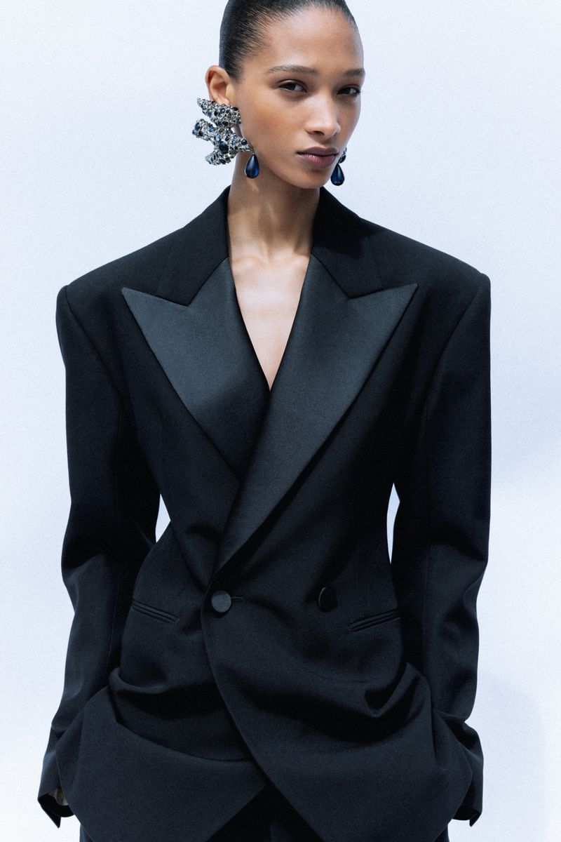 Model Elodie Guipaud wearing an oversized black tuxedo jacket with satin peak lapels and statement earrings against a light background.