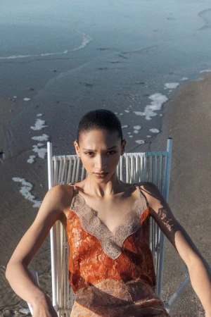 Model Elodie Guipaud seated in a white beach chair on wet sand at the shoreline wearing a rust orange lace slip dress.