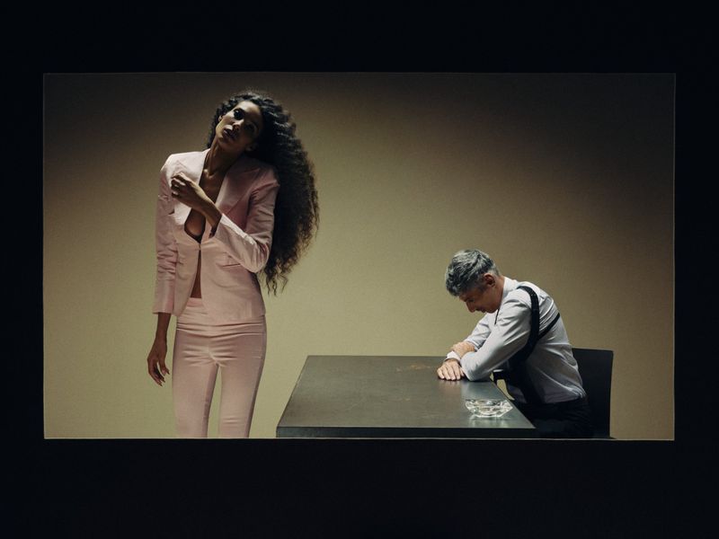 Woman in a pale pink tailored suit stands near a seated man in a white shirt and shoulder holster at a table.
