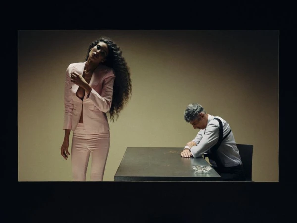 Woman in a pale pink tailored suit stands near a seated man in a white shirt and shoulder holster at a table.