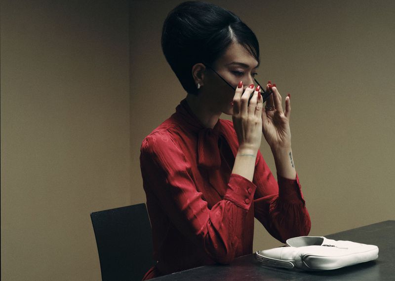 Model with a 1960s bouffant hairstyle wearing a red silk blouse while adjusting sunglasses at a table with a white purse.