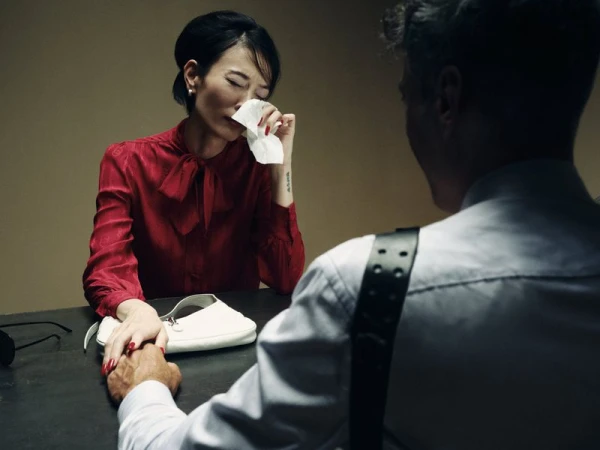 Woman in a red bow-tie blouse wiping tears with a tissue at a table, holding hands with a man wearing a shoulder holster.