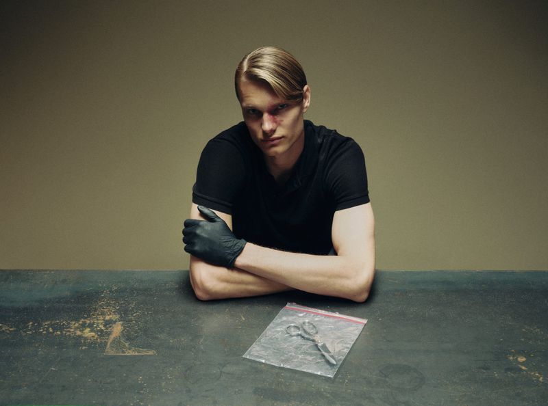 Man with a bruised nose wearing a black t-shirt and single black glove seated at a table with scissors in an evidence bag.