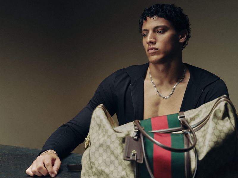 Model in an open black jacket sitting beside a monogram canvas duffle bag with a green and red web stripe against a neutral backdrop.