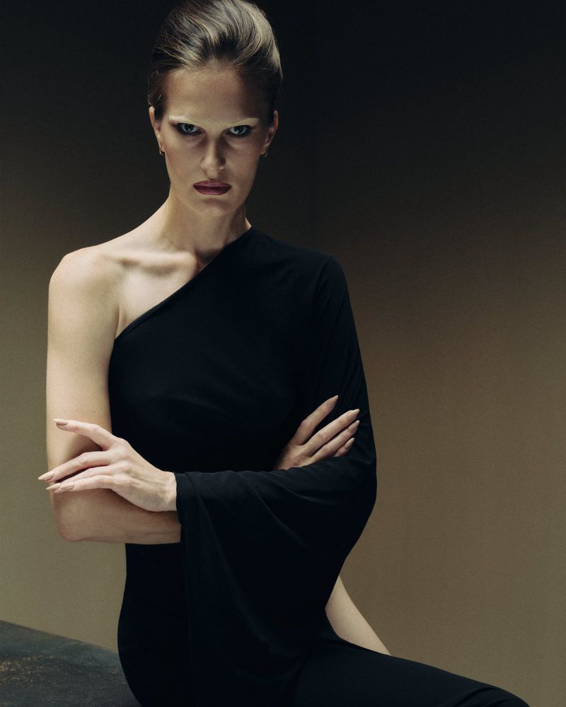 Model wearing a black one-shoulder dress with arms crossed against a dark gradient studio background