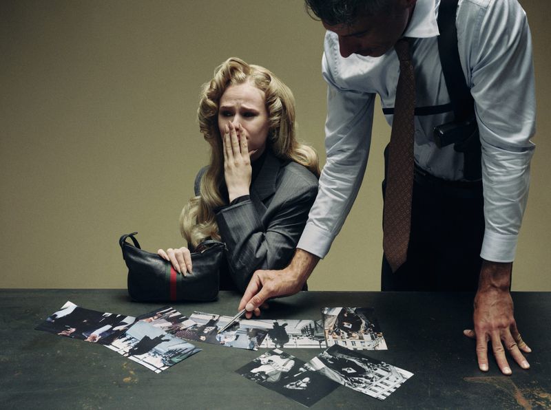 A man in a shirt and shoulder holster questions a distressed blonde woman seated at a table with photos and a black leather handbag.