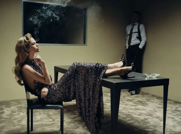 Woman in a sequined gown smoking with feet propped on a desk while a man wearing a shoulder holster stands in the background.