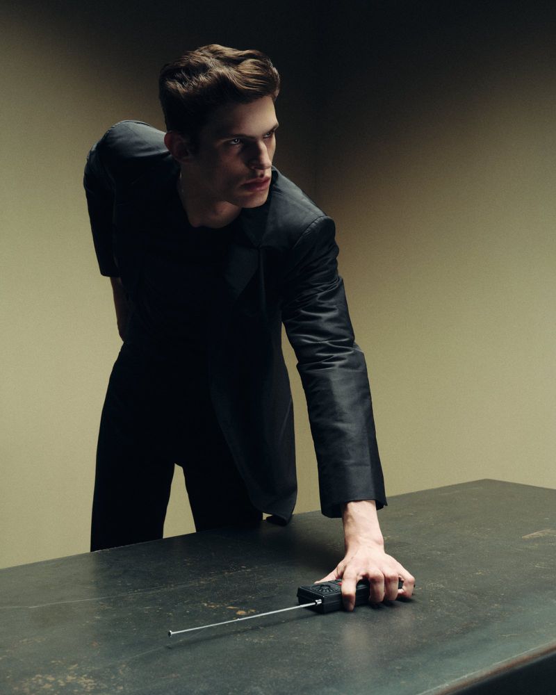 A man in a black suit leans over a dark table while touching a handheld radio with an extended antenna under dramatic studio lighting.