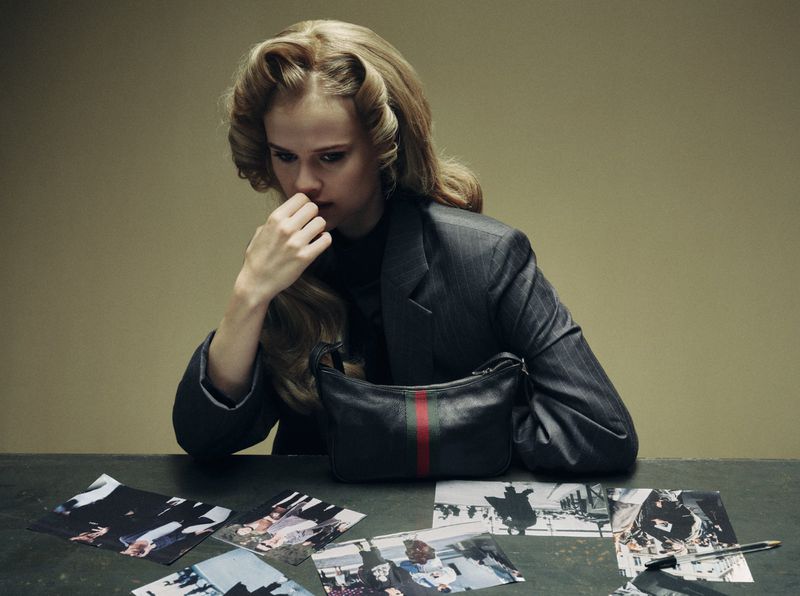Woman in a grey pinstripe jacket seated at a desk with a black leather shoulder bag and scattered photographs in a moody setting.