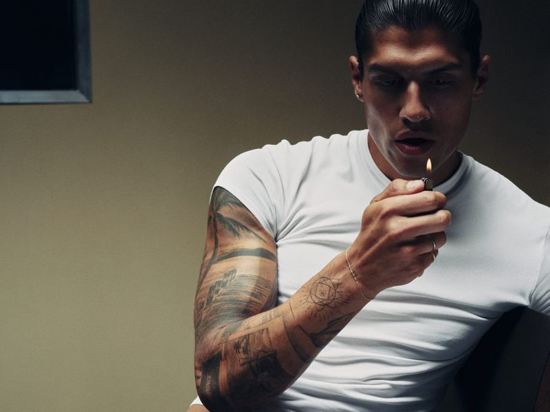 Man with full arm tattoos wearing a white t-shirt while holding a lit lighter against a muted background.