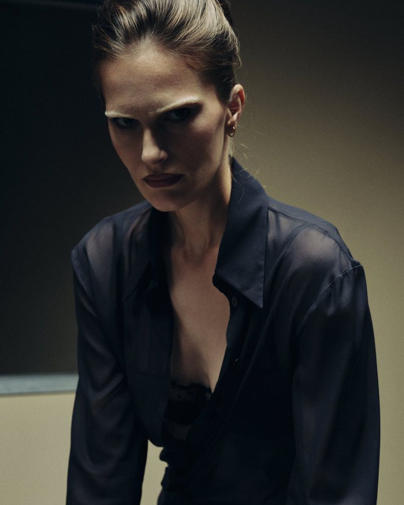Model Alla Kostromichova wearing an unbuttoned sheer black shirt looking directly forward under moody low-key studio lighting.
