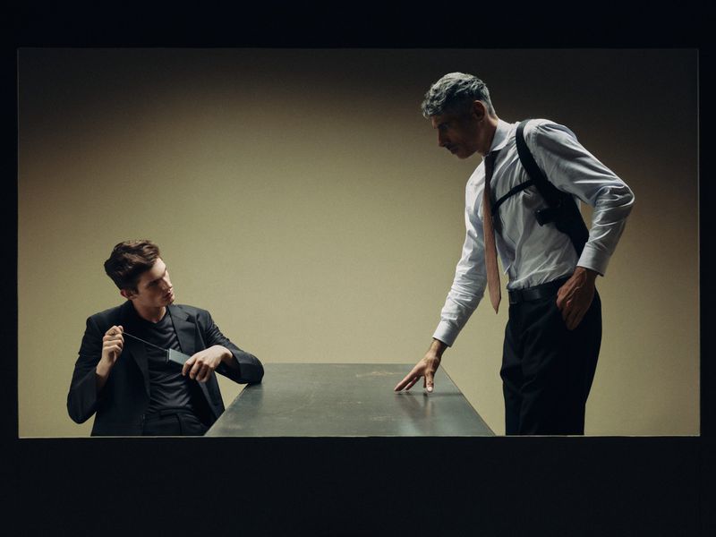 Two men in suits recreating an interrogation scene across a metal table in an editorial fashion shoot.