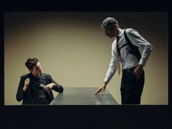 Two men in suits recreating an interrogation scene across a metal table in an editorial fashion shoot.