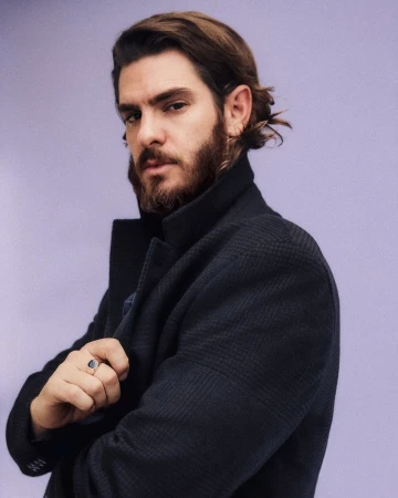 Andrew Garfield with a full beard wearing a textured dark wool coat against a lavender studio backdrop.