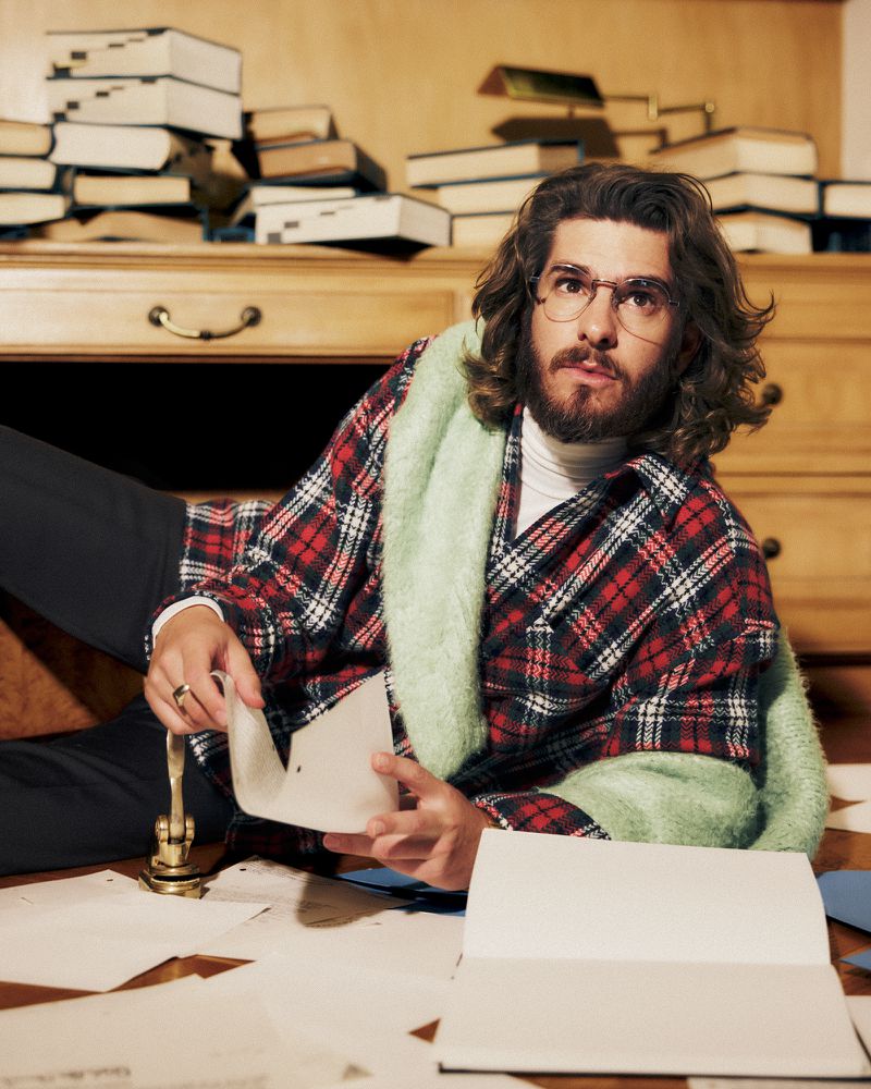 Andrew Garfield wearing a red plaid shirt, wire-rimmed glasses, and a mint green scarf lying among papers and books.
