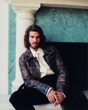 Actor Andrew Garfield seated in front of a green marble fireplace wearing a patterned brown leather jacket and cream shirt.